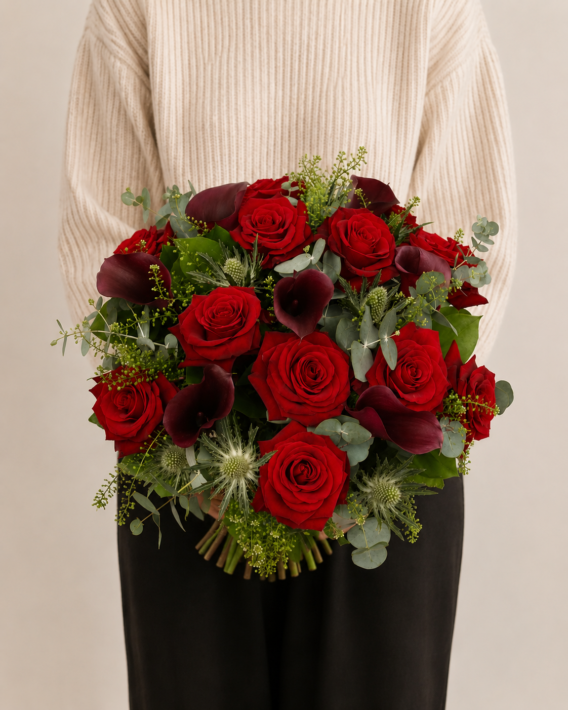 Bouquet of red and burgundy roses with greenery held by a person wearing a beige sweater.