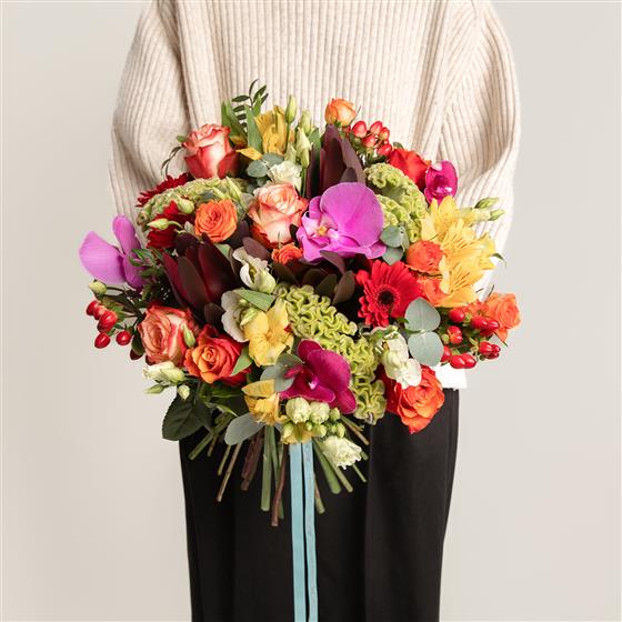Colorful bouquet held by a person wearing a light sweater and dark pants on a neutral background