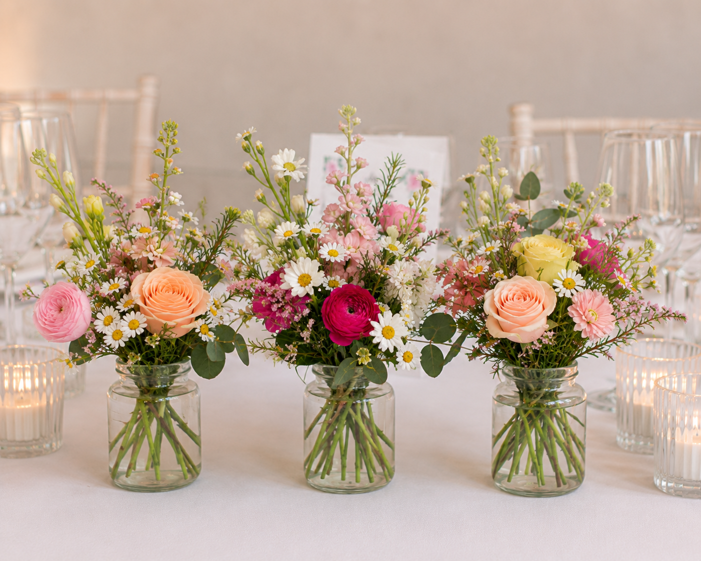 Three floral arrangements in glass jars on a table with white chairs and candles in the background.